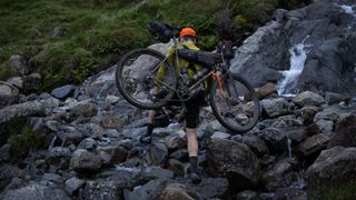 The Restrap Hike-a-Bike harness being worn as a rider carrys his bike easily over rocky stream