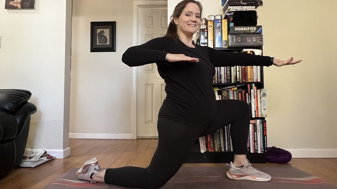 Jennifer Rizzuto performs a yoga for back mobility routine at home in her living room. She is in a half kneeling position, with her left leg bent, foot planted on the floor and thigh parallel with the ground. Her right leg is also bent, but her knee and shin lie on the ground. Her left arm is extended in front of her while her right arm is bent at the elbow; both arms are parallel to the floor.
