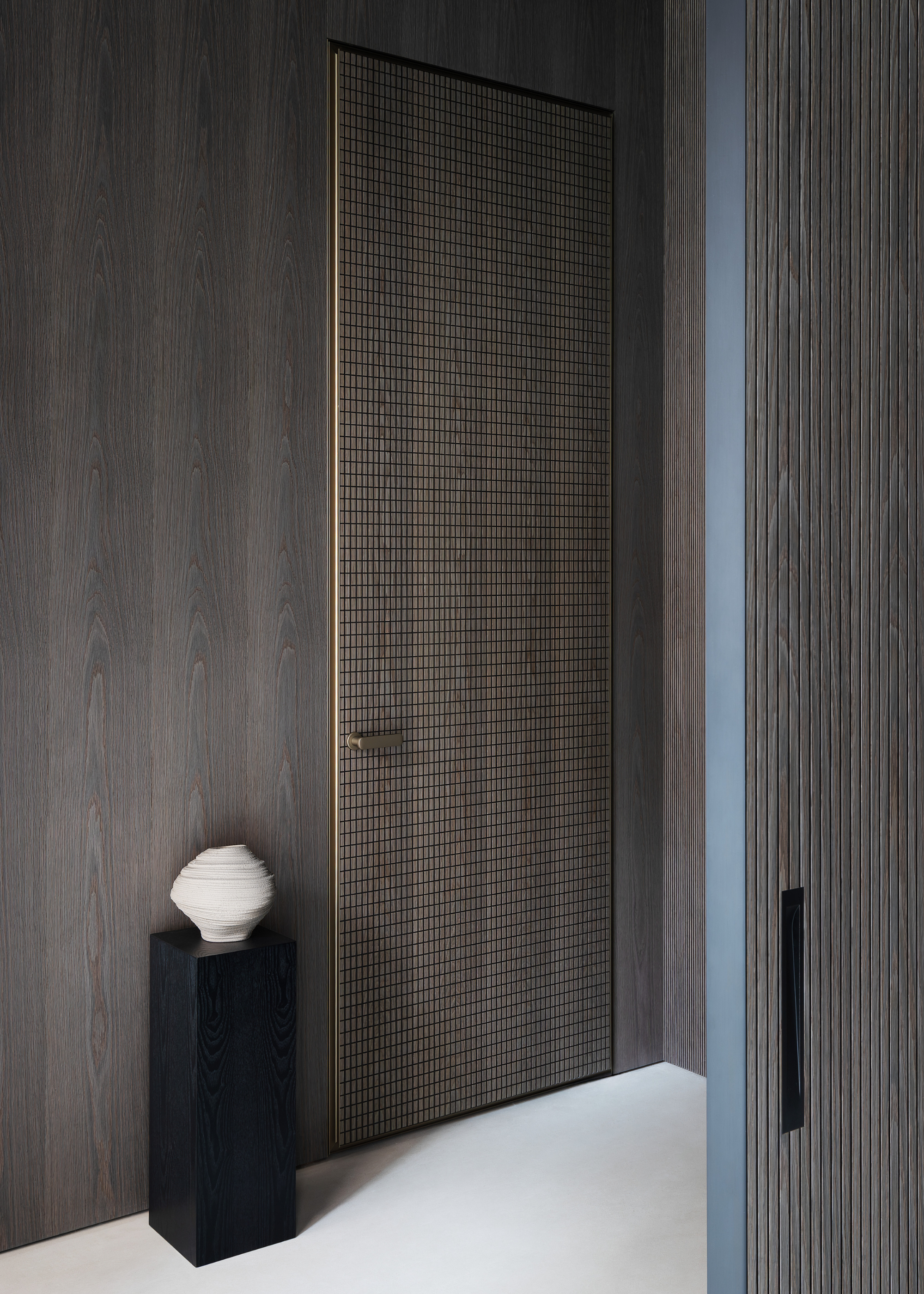 A room with a patterned wood door, wood walls, and a black plinth holding a textured white vase
