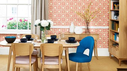 Finding out how to make a small dining room look bigger is always useful. Here is a dining room with peach and white wallpaper, a window with long gray curtains, a wooden dining table with navy blue dinnerware and wooden chairs underneath it, and a wooden sideboard and bookcase beyond it