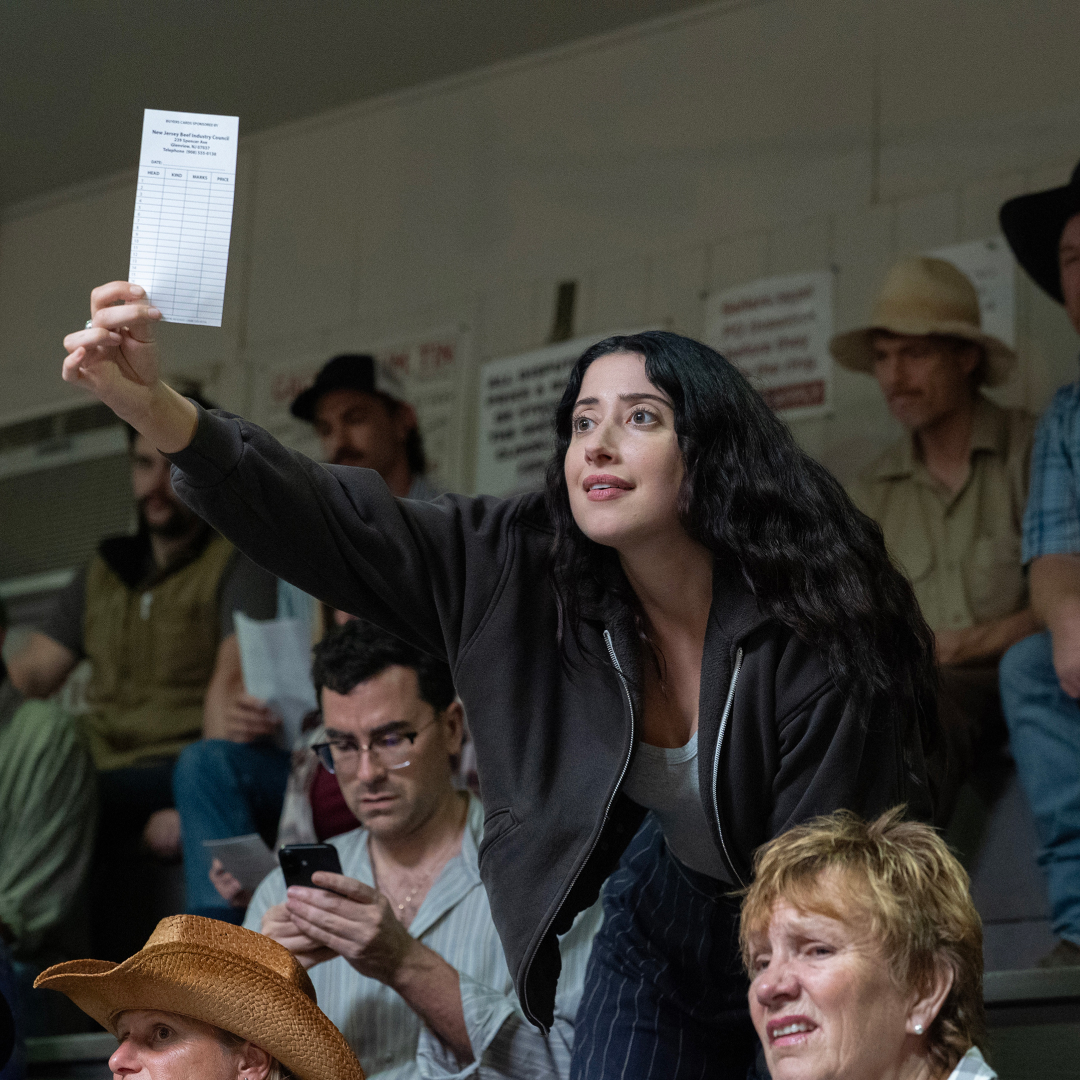 A man (Dan Levy as Nicky) looks down at his phone and a woman (Taylor Ortega as Morgan) stands while holding up a bidding slip, as they sit in a crowd at an auction, in Episode 105 of BIG MISTAKES.