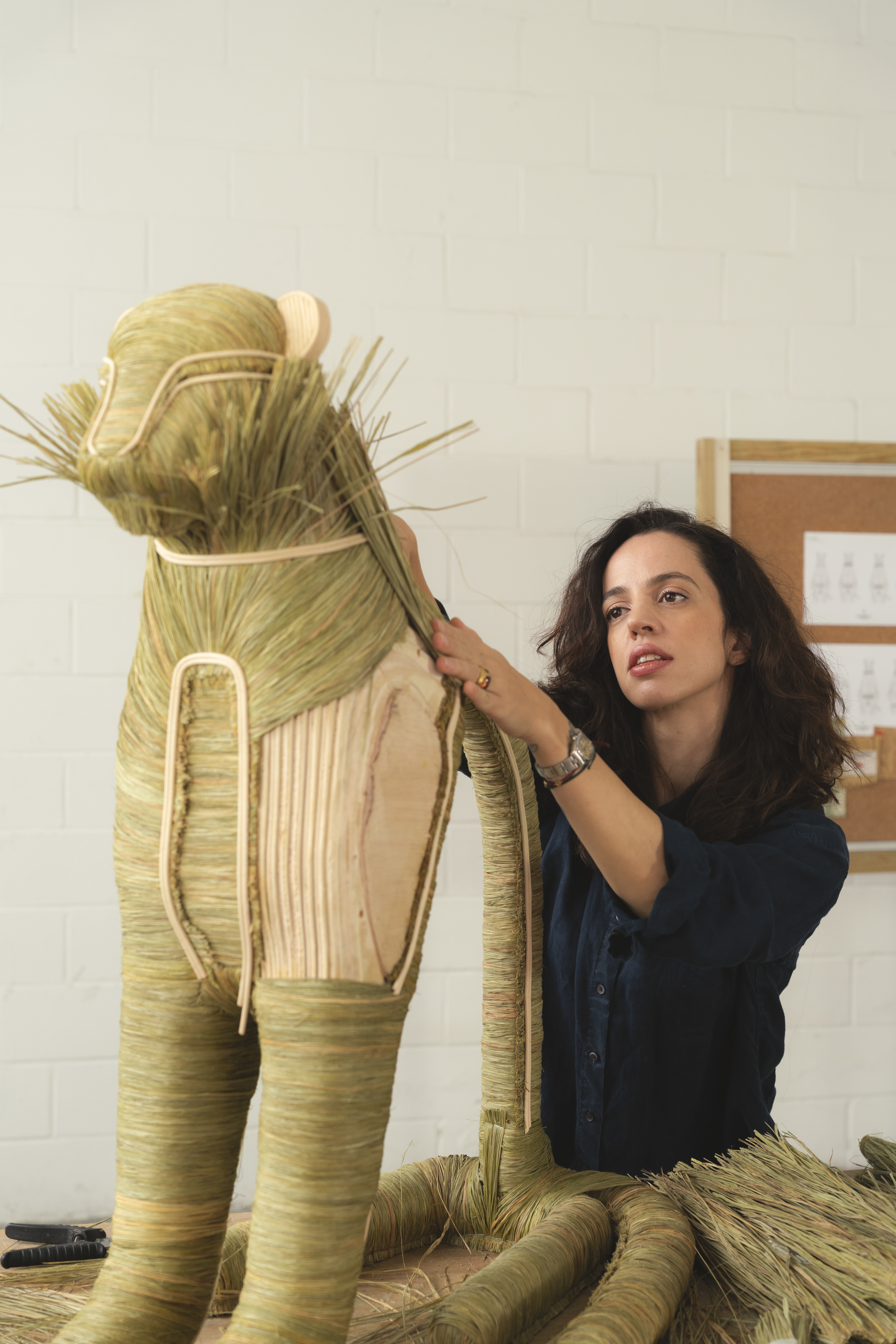 A young woman with dark long hair, wearing a dark blue buttoned-up shirt, puts the finishes touches to a tiger-like sculpture made of hay-covered wood.