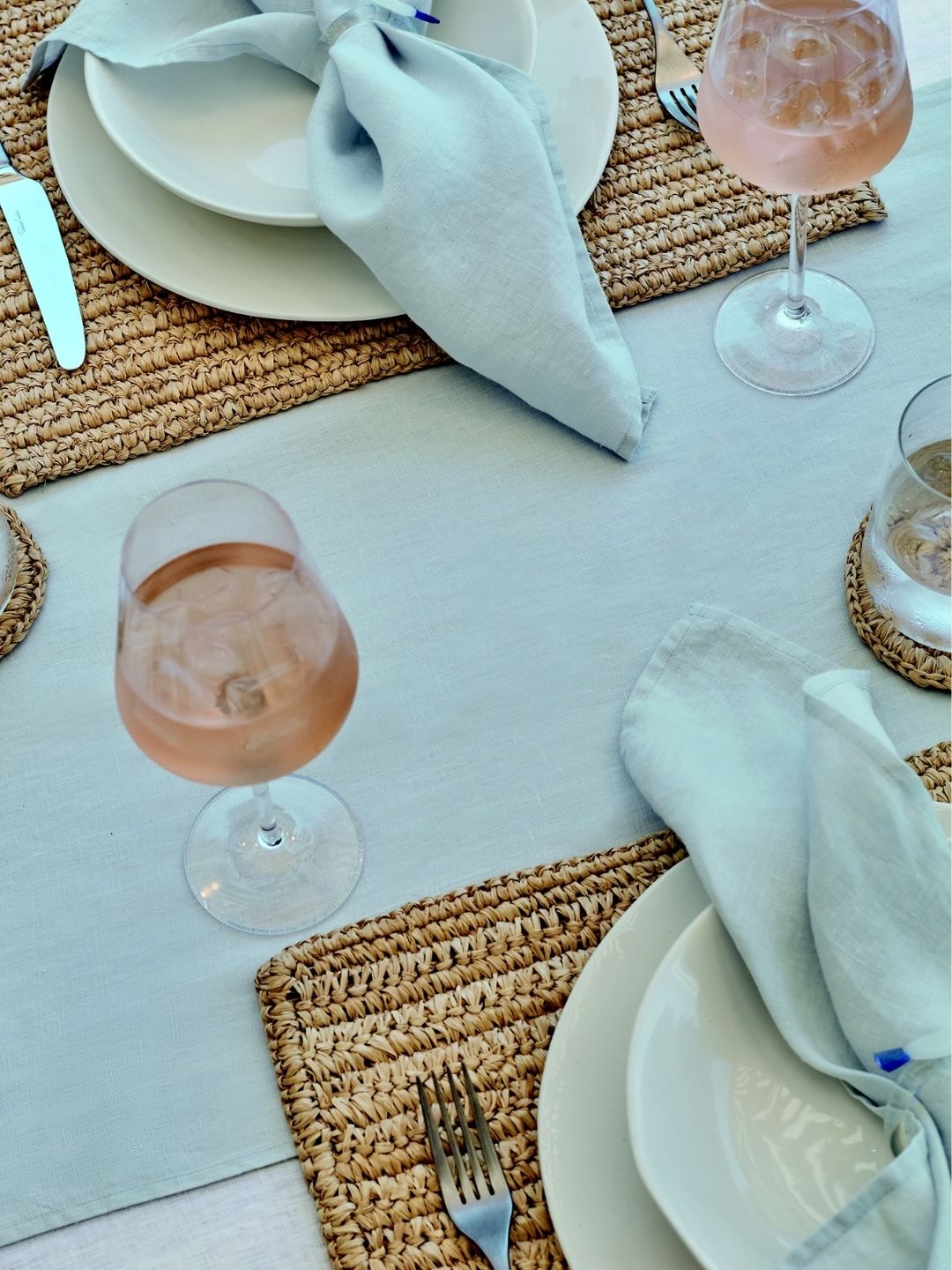 close-up image of a white table dressed with a white tablecloth, wicker placemats, white glasses, and white plates with a light blue napkin across the top.