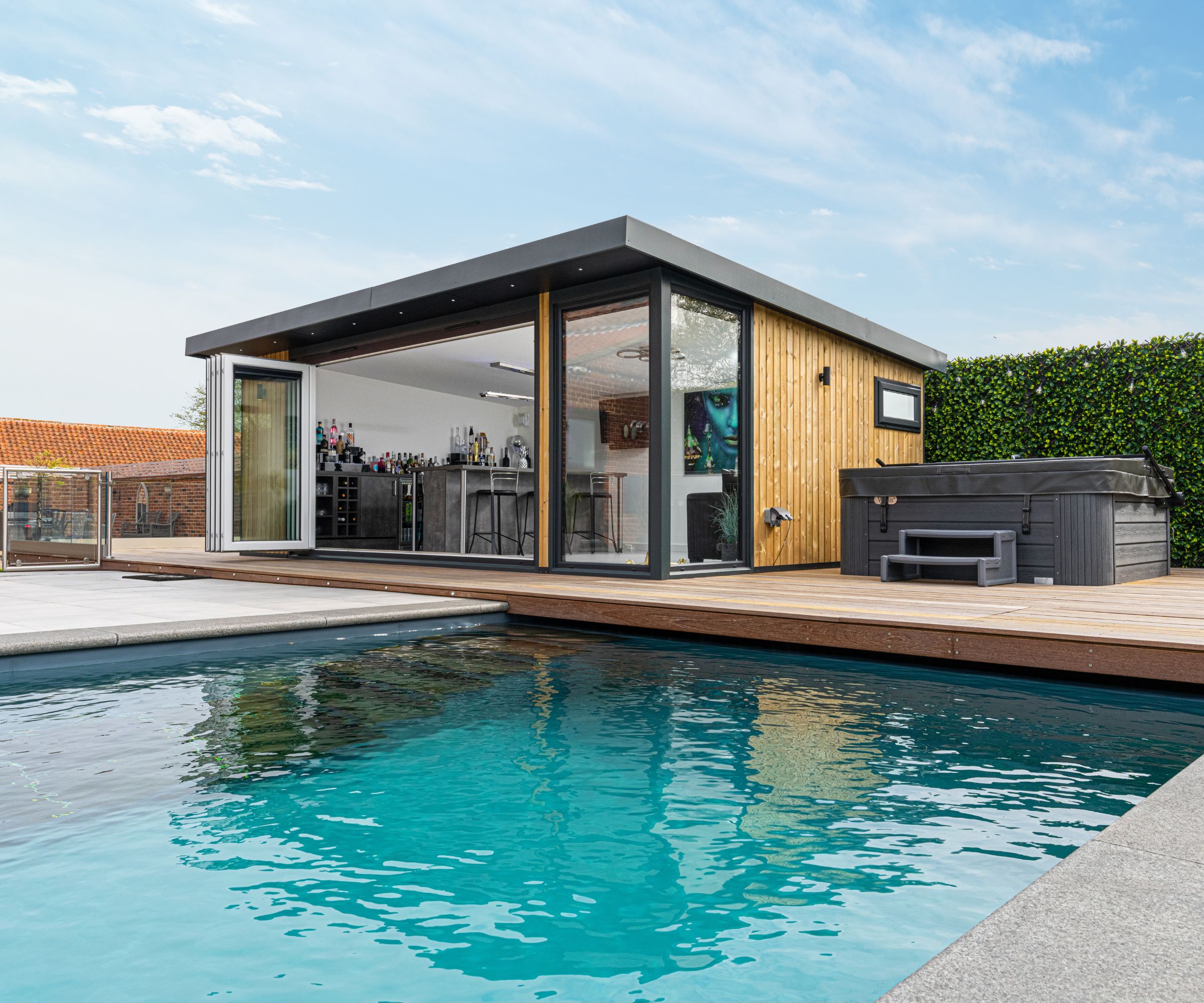 A contemporary cedar and grey flat-roof pool house with open bifold doors revealing a bar interior, a covered hot tub on the timber deck beside a turquoise swimming pool.