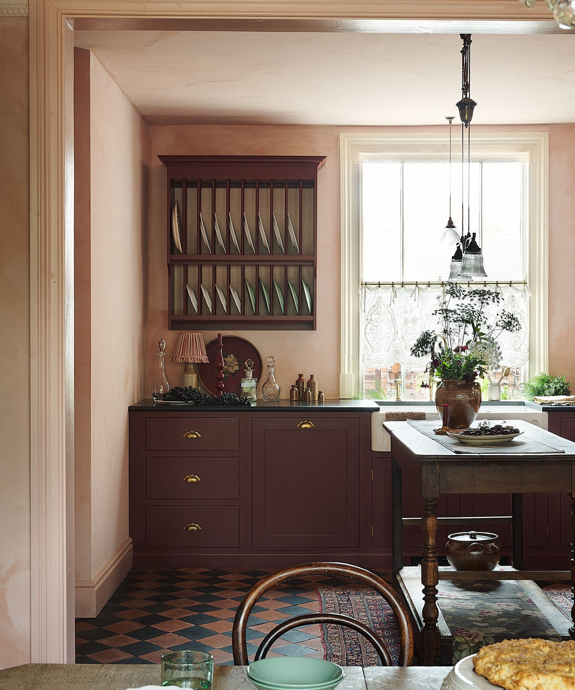 An open-plan kitchen with plaster walls, burgundy cabinets, black countertops, a wall-mounted place rack, and a wooden prep table island topped with a vase of greenery
