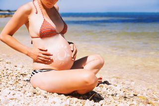 A close up of a pregant woman in a bikini putting suncream on her stomach while sat on a beach
