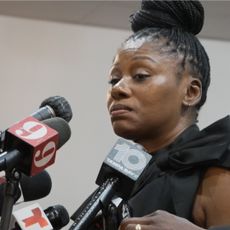 Pamela Dias speaking into several microphones at a press conference in a still from the documentary the perfect neighbor