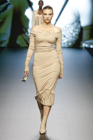 A female model wearing a form fitting long sleeved light brown dress walking down a runway.