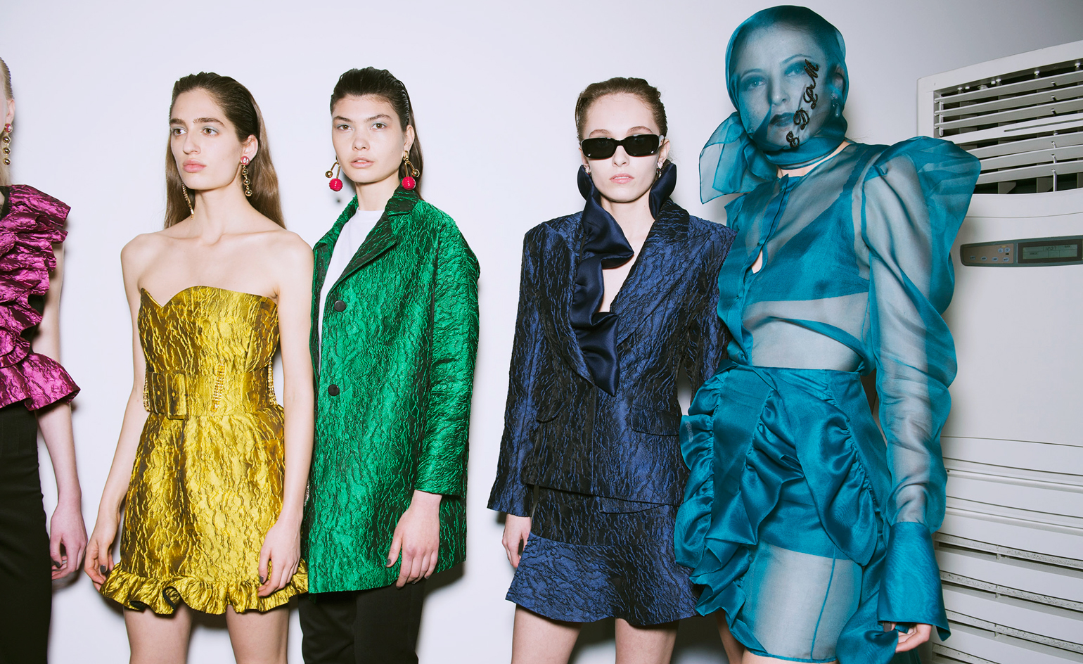 Models wear yellow, green, navy and bright blue dresses and tailored suits. One model wears a blue mesh face mask