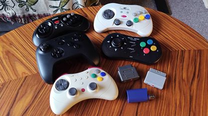 Selection of retro controllers tested by GamesRadar+ editor Phil Hayton on woodgrain table, including Retro-Bit Genesis wireless controller, Retro-Bit Sega Saturn Pro, 8Bitdo N64 controller, 8bitDo Neo Geo controller, Gulikit Elves 2 Pro, and 8BitDo Retro receivers. 