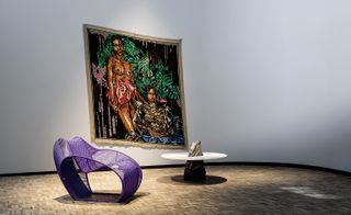 View of colourful canvas art featuring two people hanging on the wall along with a curved purple chair and a round table with a rock-like base in a space with white walls and brown patterned flooring