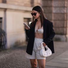 Bella Ema seen wearing a black sunglasses, silver earrings, a black oversize LeGer blazer, a creme satin crop top from LeGer, a creme linen shorts from LeGer, a white leather handbag on March 28, 2022 in Berlin, Germany