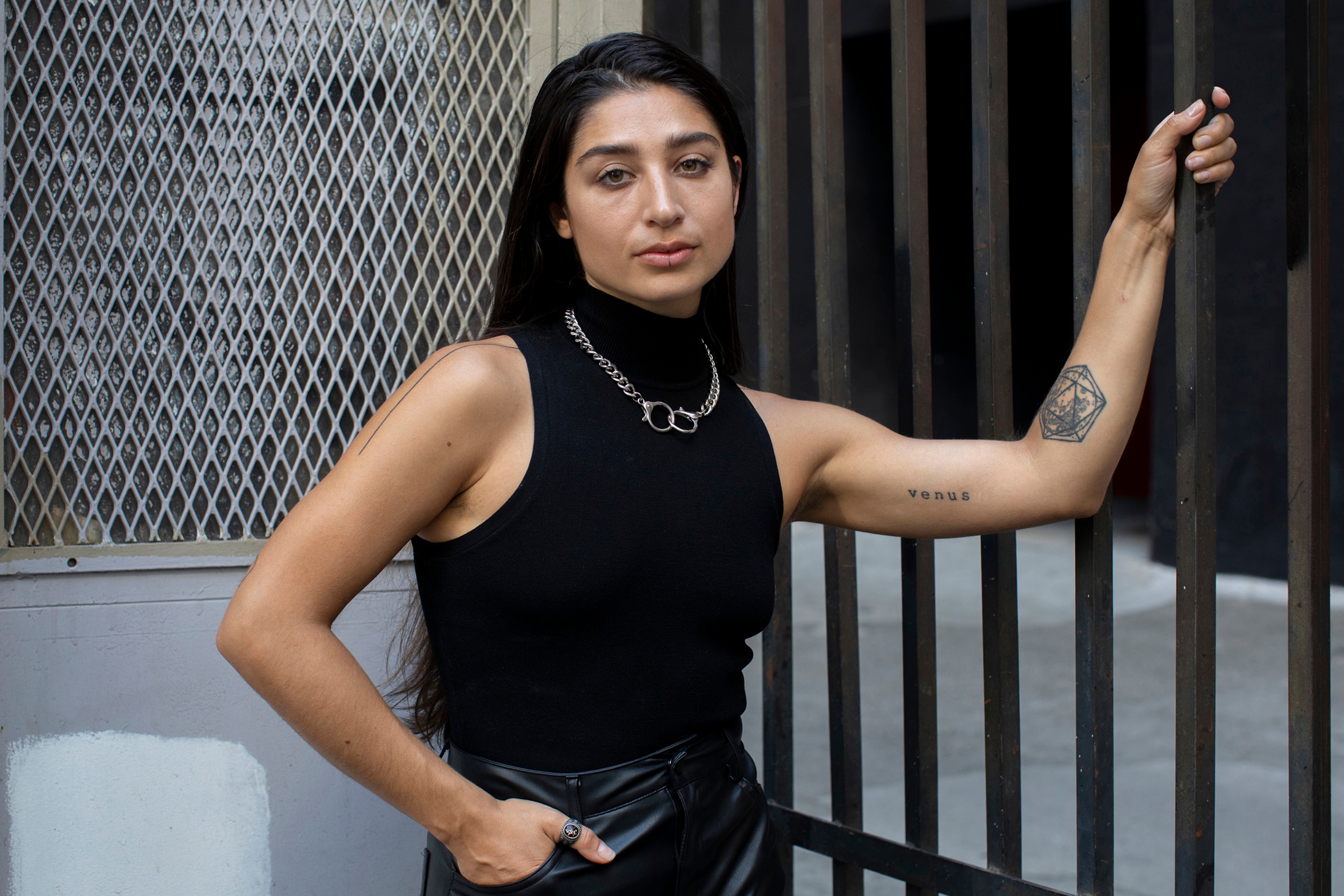 A young woman with long dark hair leans against a metal gate, wearing a black sleeveless turtleneck and leather trousers, with a geometric tattoo and the word "venus" tattooed on her forearm.
