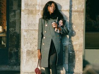 French woman wears a J. Crew coat, tights, skirt and bag while holding a magazine and drinking a coffee