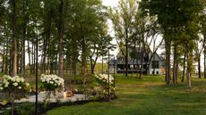 A wide shot of a modern, multi-story farmhouse-style home nestled behind a grove of tall trees, featuring a manicured green lawn and white flowering hydrangeas in the foreground.