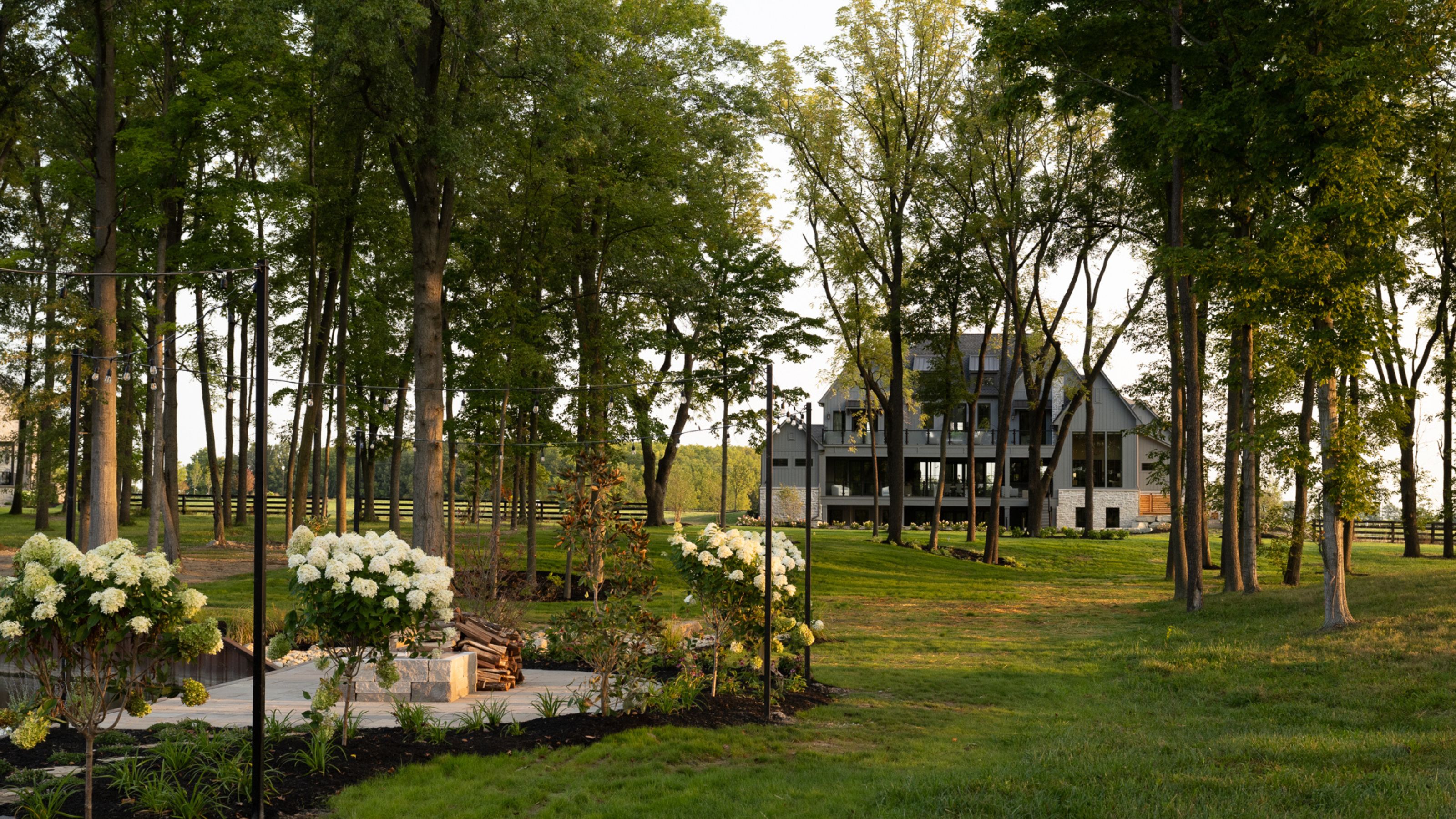 A wide shot of a modern, multi-story farmhouse-style home nestled behind a grove of tall trees, featuring a manicured green lawn and white flowering hydrangeas in the foreground.