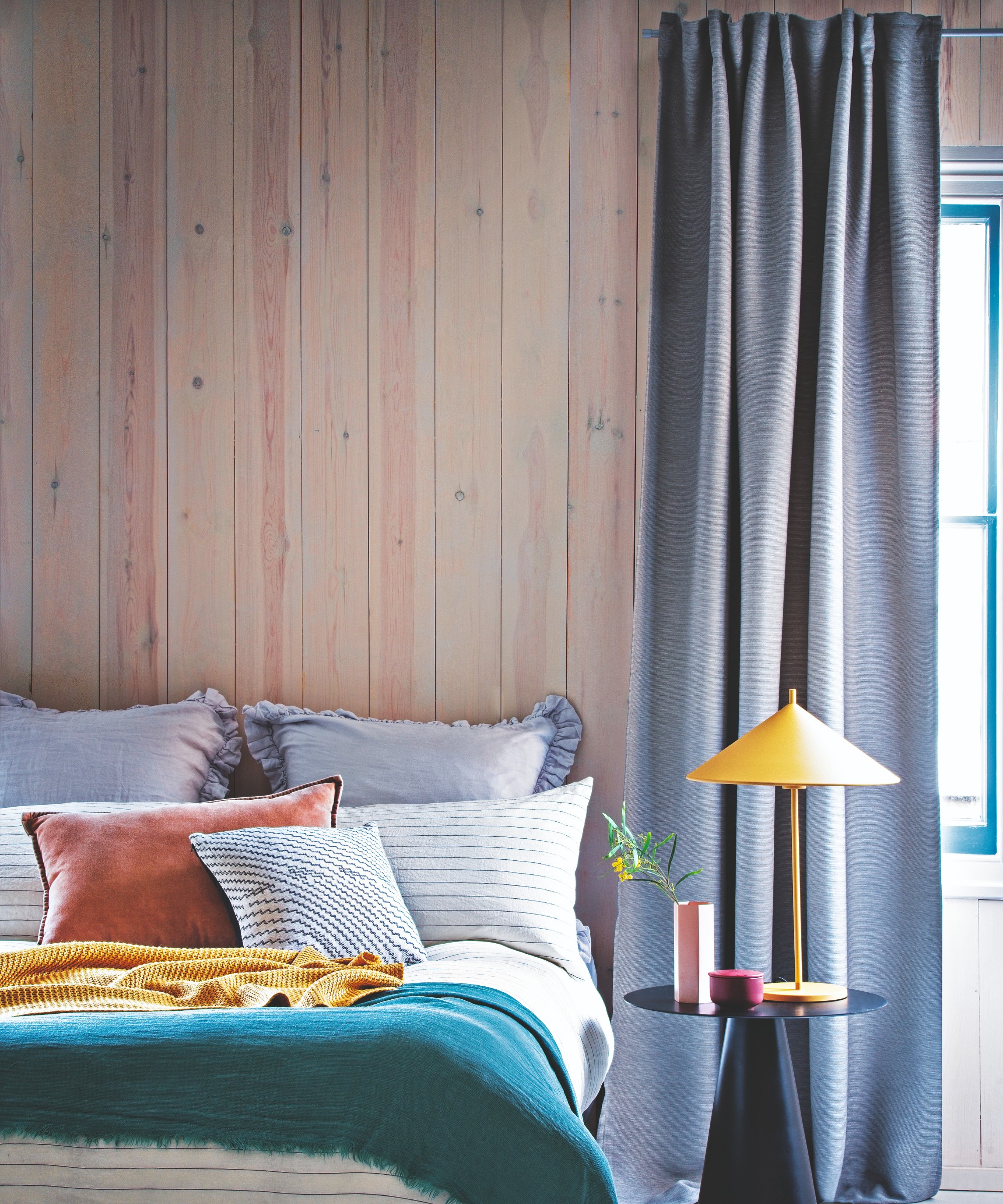 A bedroom with panelled walls and steel blue curtains against a bright yellow table lamp on the bedside table