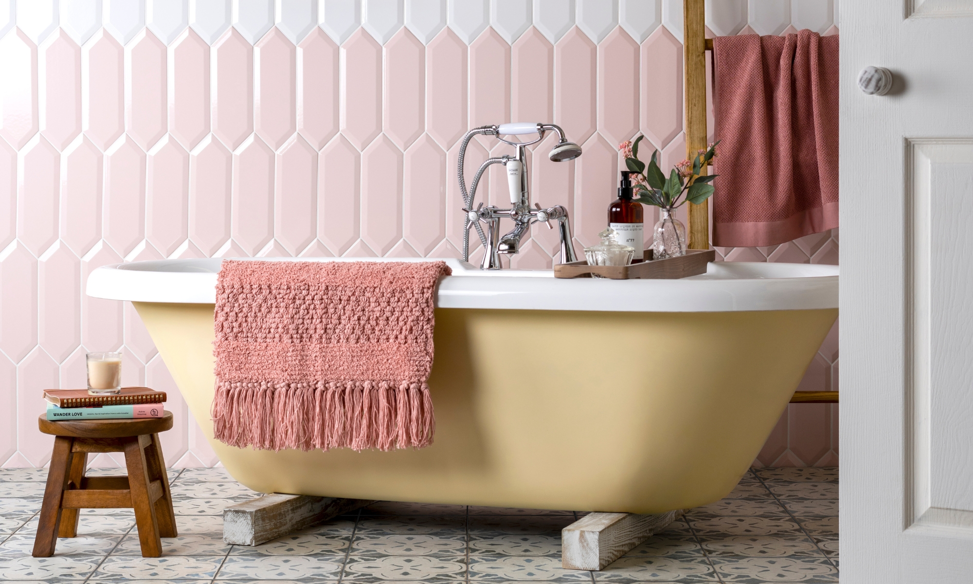 Yellow roll-top bath with pink towel against pink and white geometric tile, patterned floor tiles.