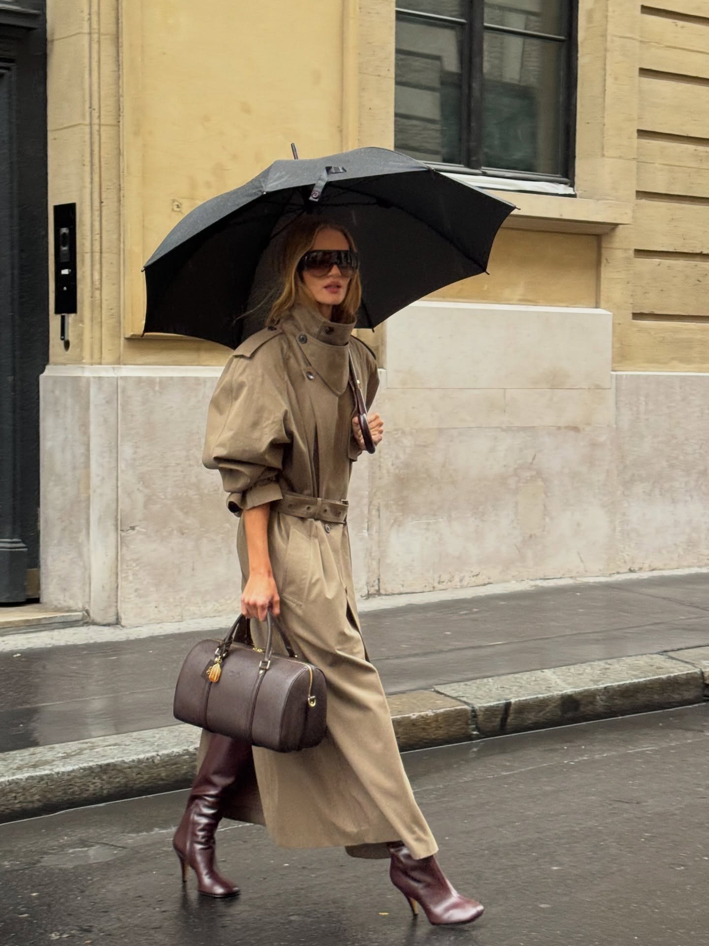 Model Rosie Huntington-Whiteley walks outside in the rain underneath an umbrella wearing a beige funnel neck trench coat with burgundy boots and a bowling bag.