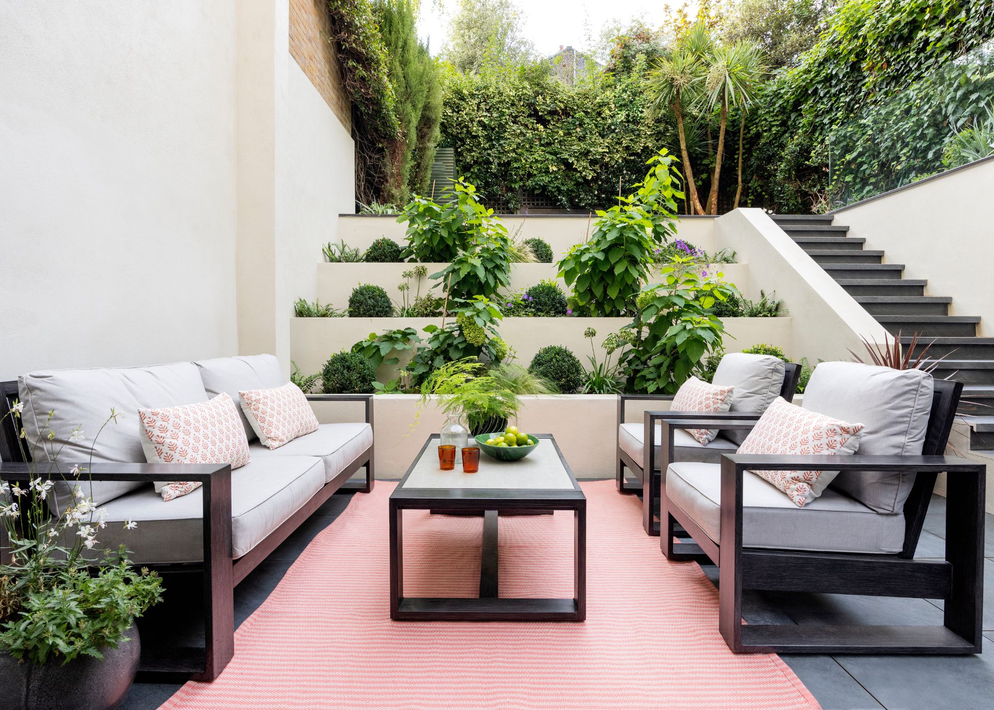 A modern garden with a white exterior, a patio seating area with a gray minimalist outdoor sofa and two matching chairs. A red rug, a coffee table, and steps with levels of lush green planting.
