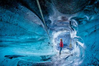 a female tourist wearing red with helmet standing at the opening of an ice cave in Iceland
