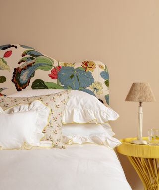 bed with white ruffle bedding, floral upholstered headboard and yellow side table