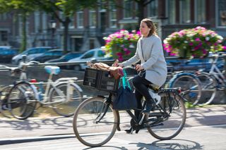 Should I cycle with earphones? This image shows a woman riding a bike in non-cycle wear, with a basket on the front of her bike, she is wearing headphones. In the back ground are lots of bikes lent against railings which have flowers in baskets on them.