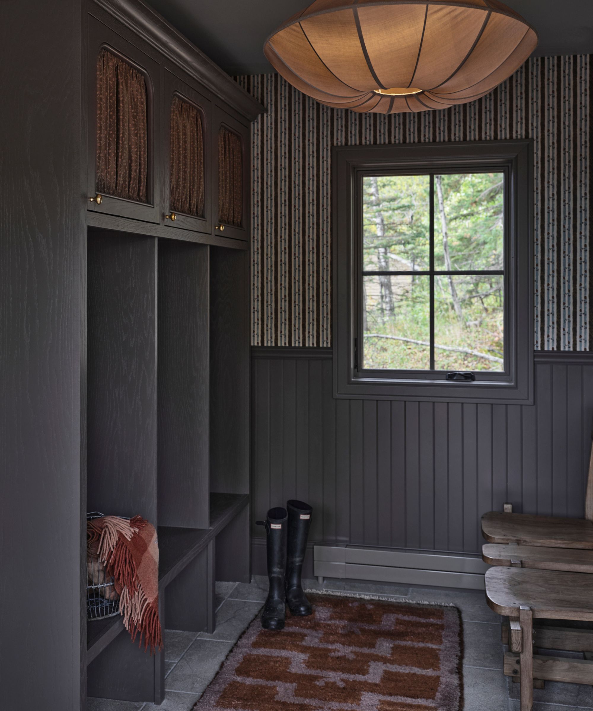 a warm mauve entryway with floral striped wallpaper, half height paneling, limestone floors, a vintage stool an drug and silk pendant light