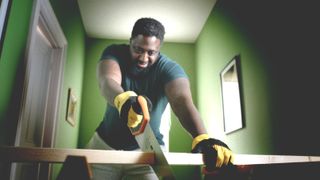 man sawing length of wood wearing goggles in room with green painted walls