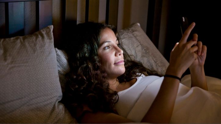 A woman with brown curly hair in bed on her phone as she battles phone addcition