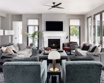 Grey painted living room with large seating space, two sofas and three armchairs gathered around a central coffee table, white fireplace with tv mounted above on the wall, rich with texture and subtle color palette