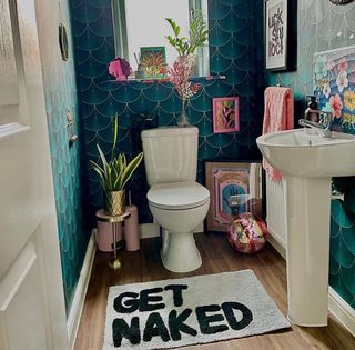 A bathroom with teal tiled walls and a bath mat that says 'get naked'