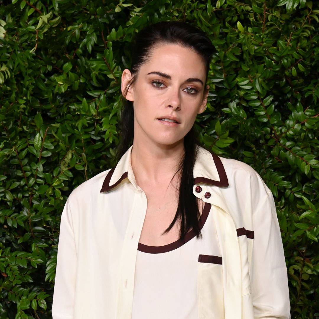 Kristen Stewart wearing a half up half down hairstyle. She is wearing a collared white shirt with a black trip and is standing in front of a bush. 