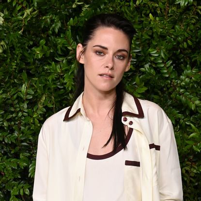 Kristen Stewart wearing a half up half down hairstyle. She is wearing a collared white shirt with a black trip and is standing in front of a bush. 