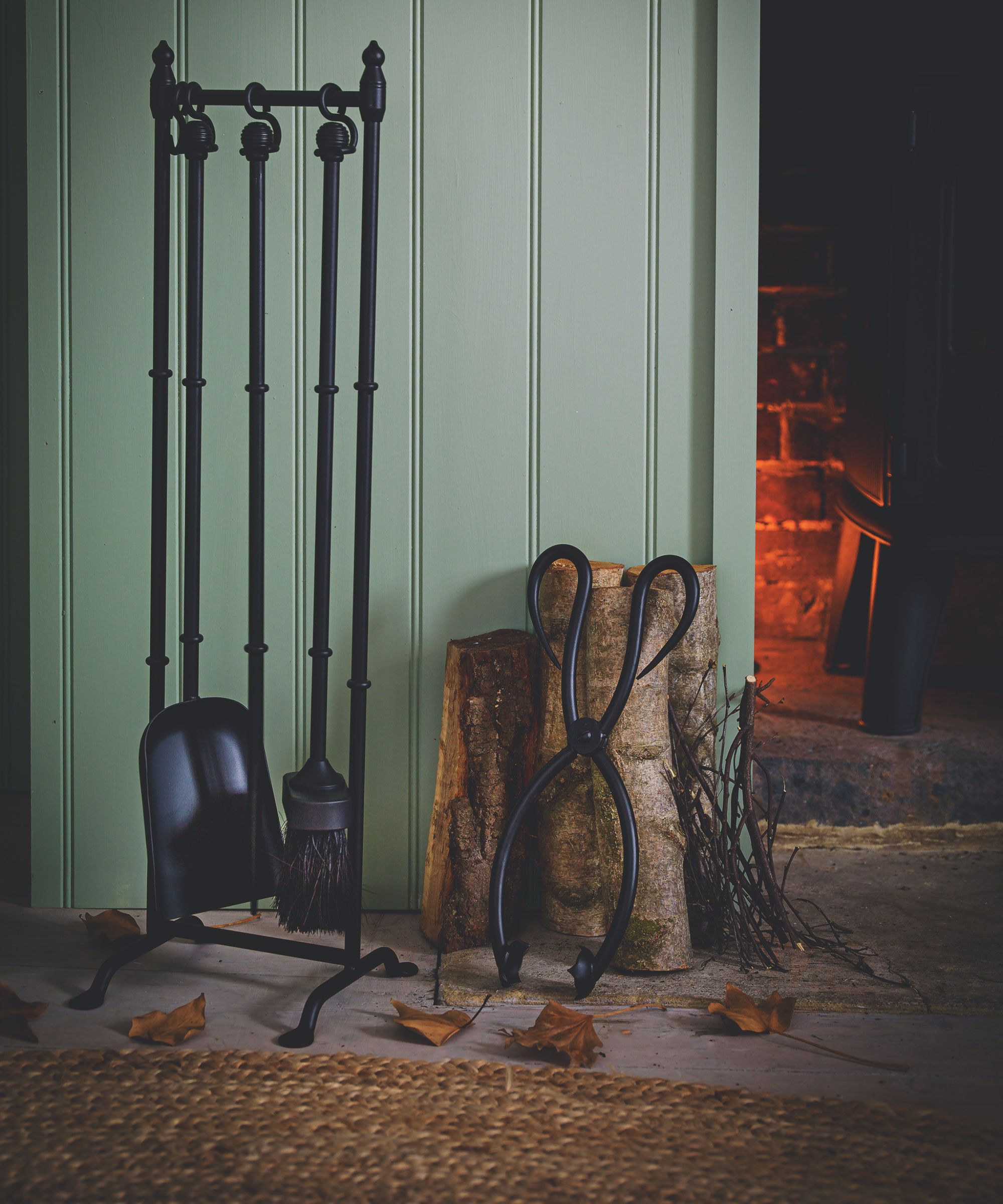 A green panelled wall next to an exposed brick fireplace with a lit fire. In front of the panelled wall are logs of wood and various black metal fire accessories hanging from a rail, with a rattan rug.