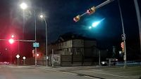 a bright flash of light crosses the night sky above businesses and street lights along a road