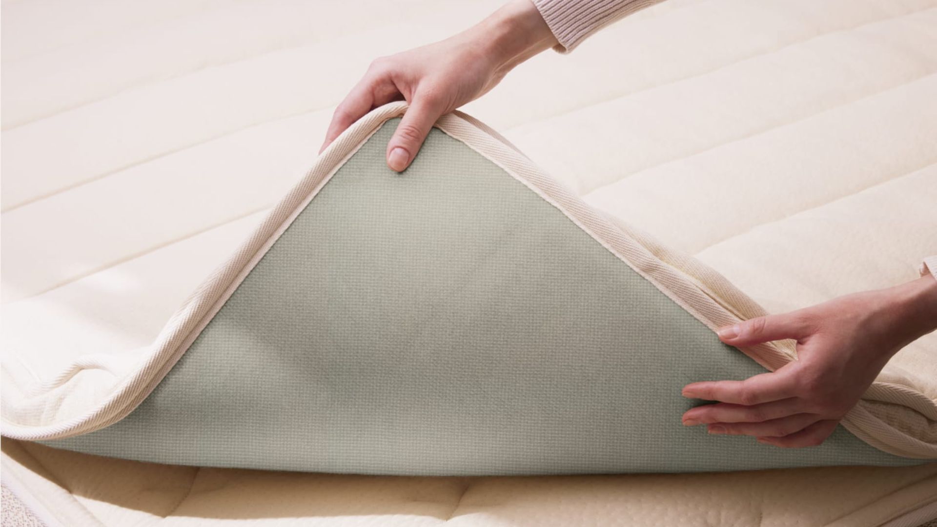 A woman&#039;s hands lifts the corner of the Birch Plush Organic Mattress Topper to reveal its underside.