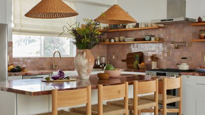 A warm kitchen with blush zellige tiles on the backsplash, floating wood shelves, rattan style pendants, a terracotta vase, a chopping board with fruits and vegetables, and a set of four woven wood stools