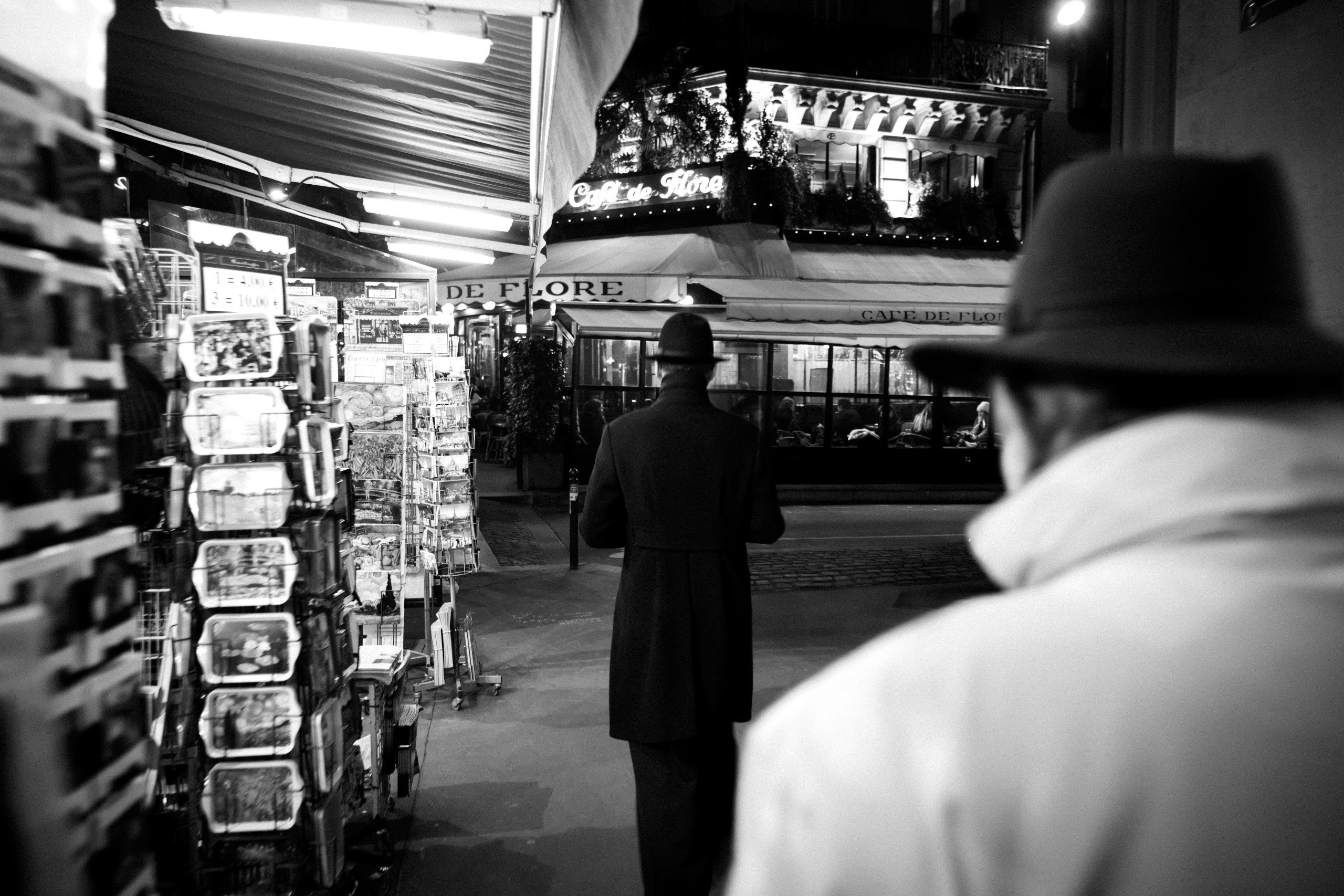 A man in a long coat and bowler hat walks alone at night past the illuminated awning of Caf&amp;eacute; de Flore, seen past a rack of postcards.