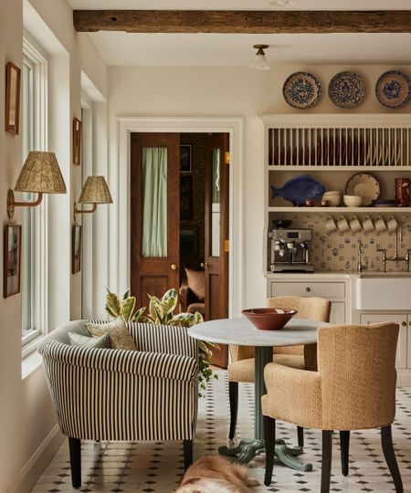 The corner of a warm neutral kitchen with a living-room style seating area, complete with a striped love seat, two yellow upholstered chairs, and a round white tabletop on a green pillar-style stand