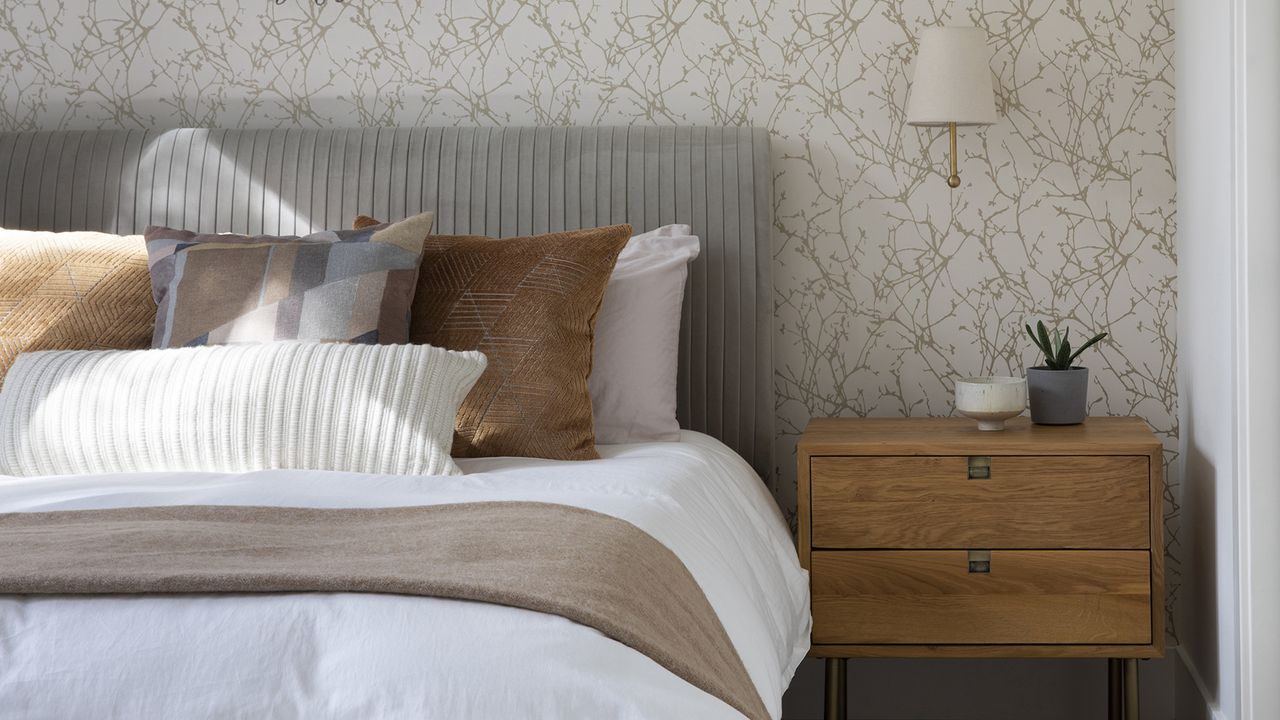 Guest bedroom with bed with white linen, neutral pillows, nightstand, wall light, and patterned neutral wallpaper