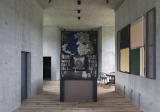 A kitchen area with a black island counter, high roof, concrete walls, wooden floor and a large book shelf.