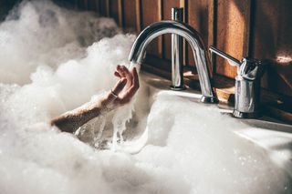 The image shows a woman's hand emerging from a hot bath to run under the cold tap