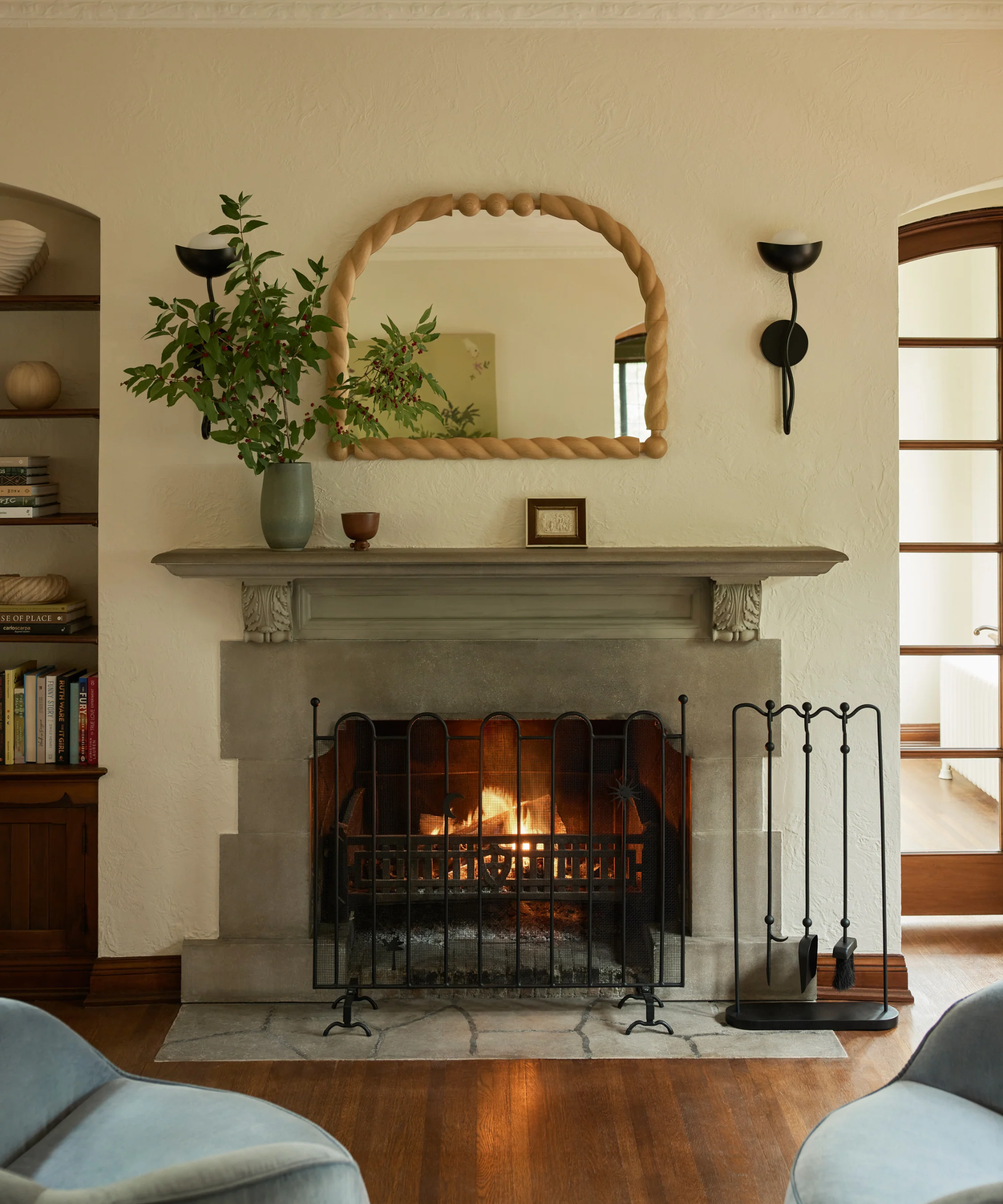 a living room with a stone fireplace, a metal fireplace screen, a wooden mirror, and a plant in a vase on the mantel