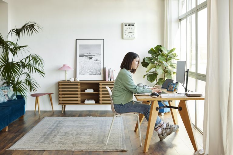 Side view of busy female entrepreneur using computer at home office, plants for an office desk. Businesswoman is working while sitting on chair at table. She is wearing casuals.