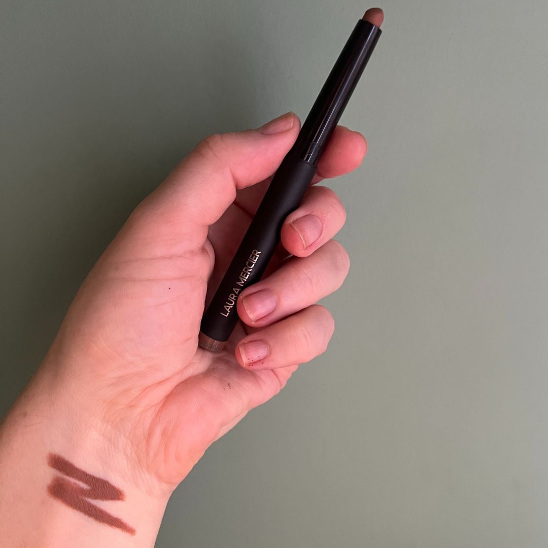 a hand holding laura mercier cavier eye stick with a scribble of the eye crayon on the wrist against a green backdrop