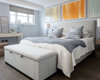 A pale blue bedroom with storage bench and orange artwork