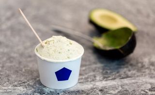 a white tub with blue hexagon on it filled with green speckled ice-cream and a wooden spoon on a grey counter next to an avocado skin and spoon