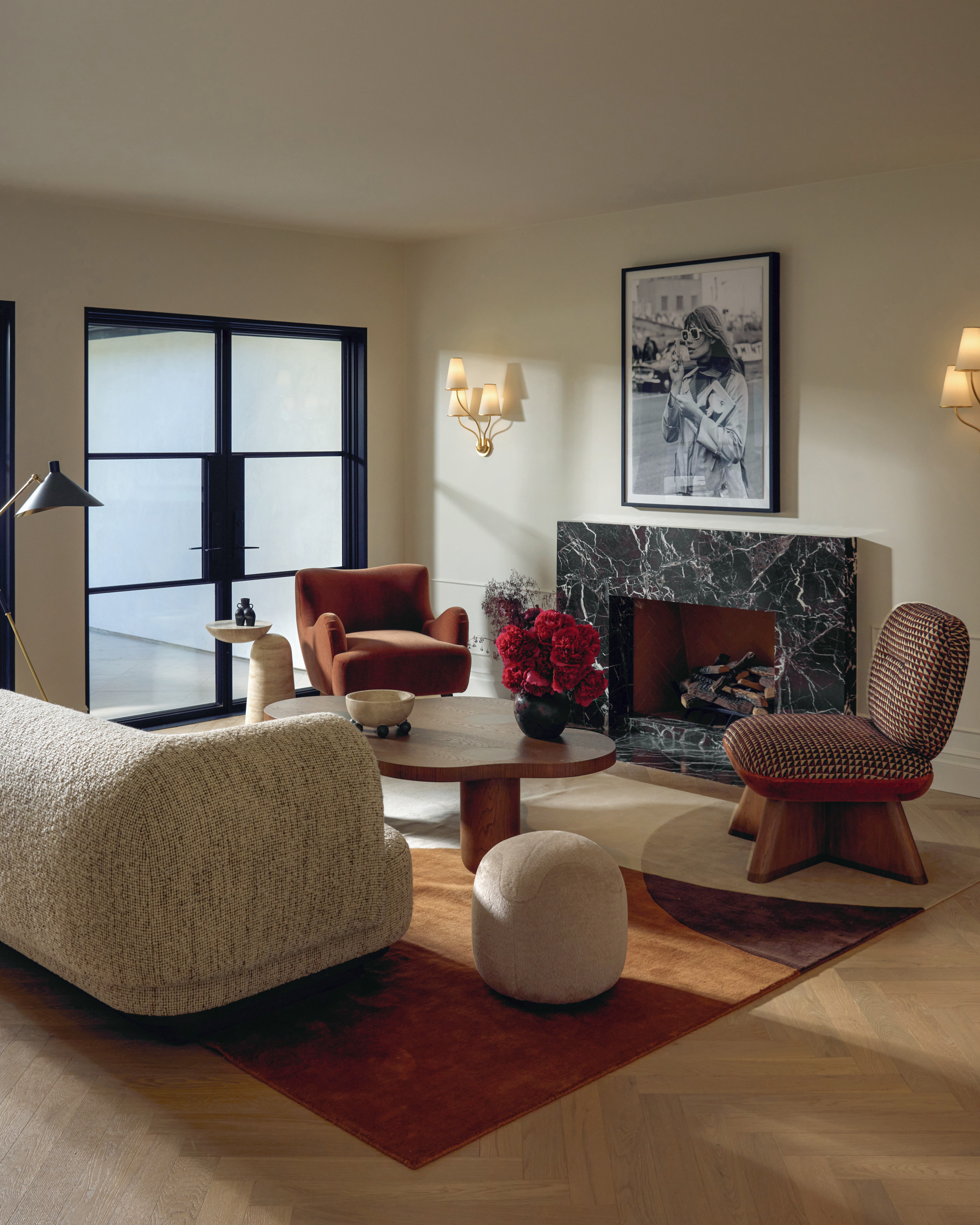 Cream living room with black marble fireplace, multi-bulb wall sconces either side framing a photograph of Fran&amp;ccedil;oise Hardy, cream sofa, cream pouffe, wood wavy cofee table, wood and burgundy fabric armchairs and rust colored rug