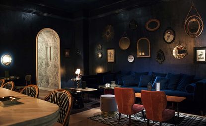 Dark theme with couches and low single seater chairs with old picture frames and mirrors hanging from the wall